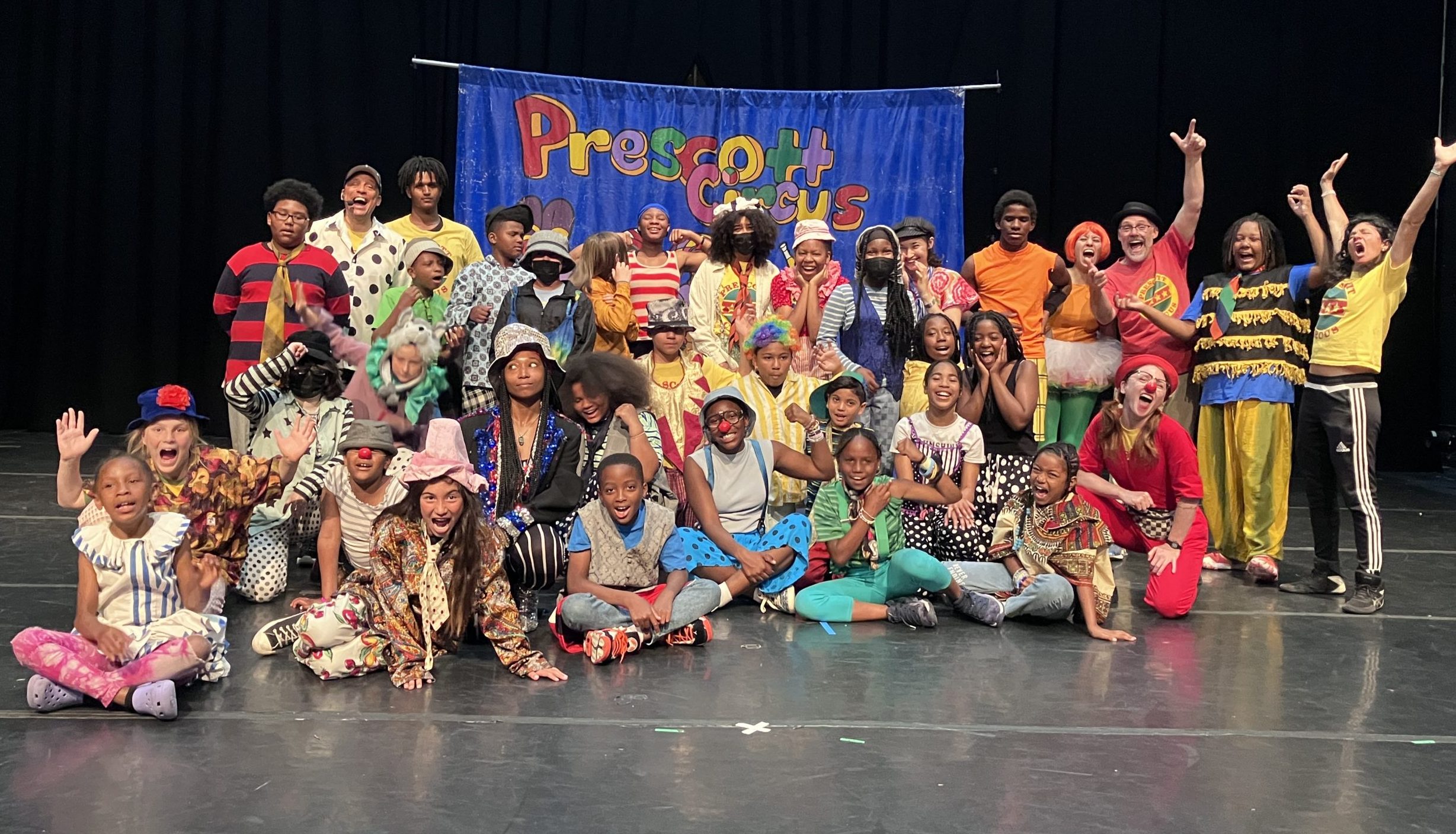 summer 2023 performing company. A group of children with supporting adults are posing in a group dressed in colorful costumes smiling.