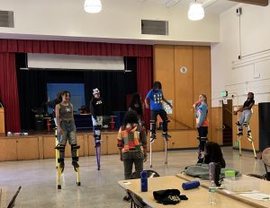 Six students are practicing dancing on stilts in the auditorium. Coach coicoi is standing in front of them with her back to the camera giving them instruction.