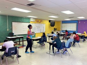 Miss Sparkle is walking around the classroom helping students sitting at their desks working on projects. 