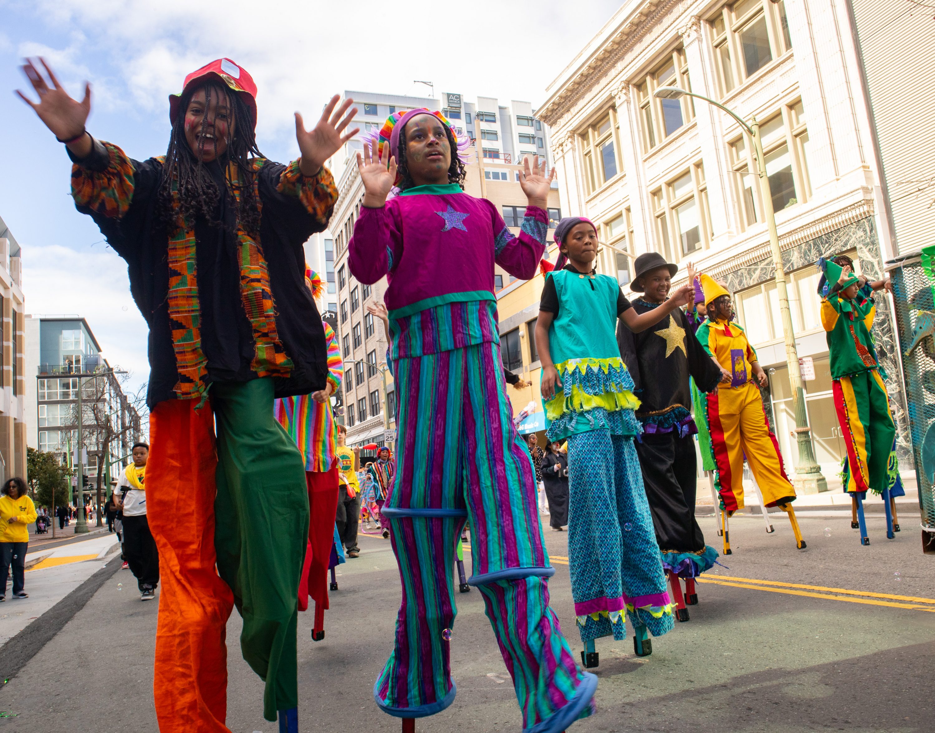 Five young stilt walkers, some wearing African print costumes, wave and smile walking down the street.