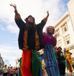 Two stilt walkers are standing tall in front of blue sky and white building. One has their arms raised up and is wearing African print costume. The other is making peace signs with their hands and wearing color circus costumes.