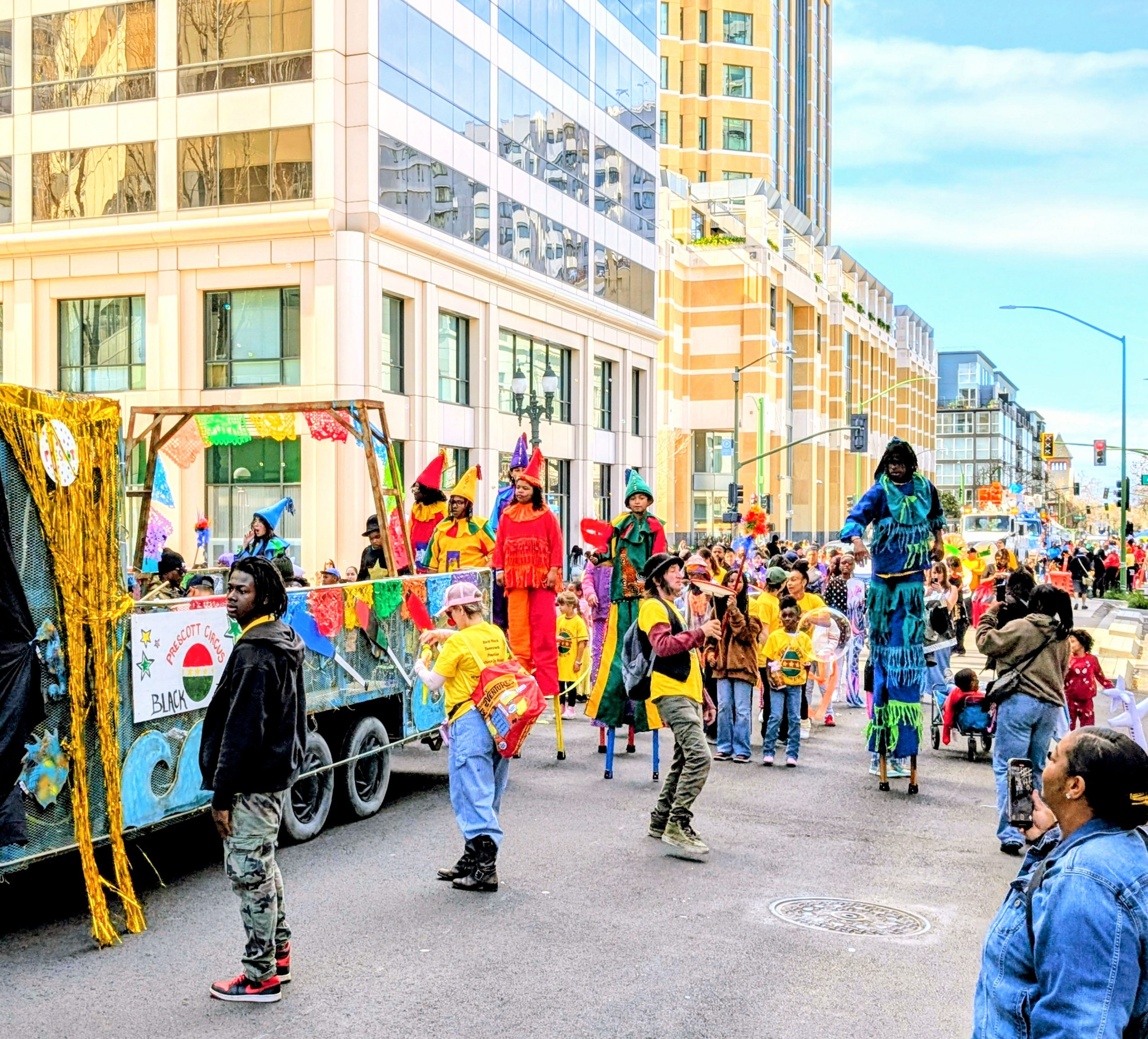 Many circus students on stilts and on the ground in costume stand on the street behind The Pacific art car for the Black Joy Parade