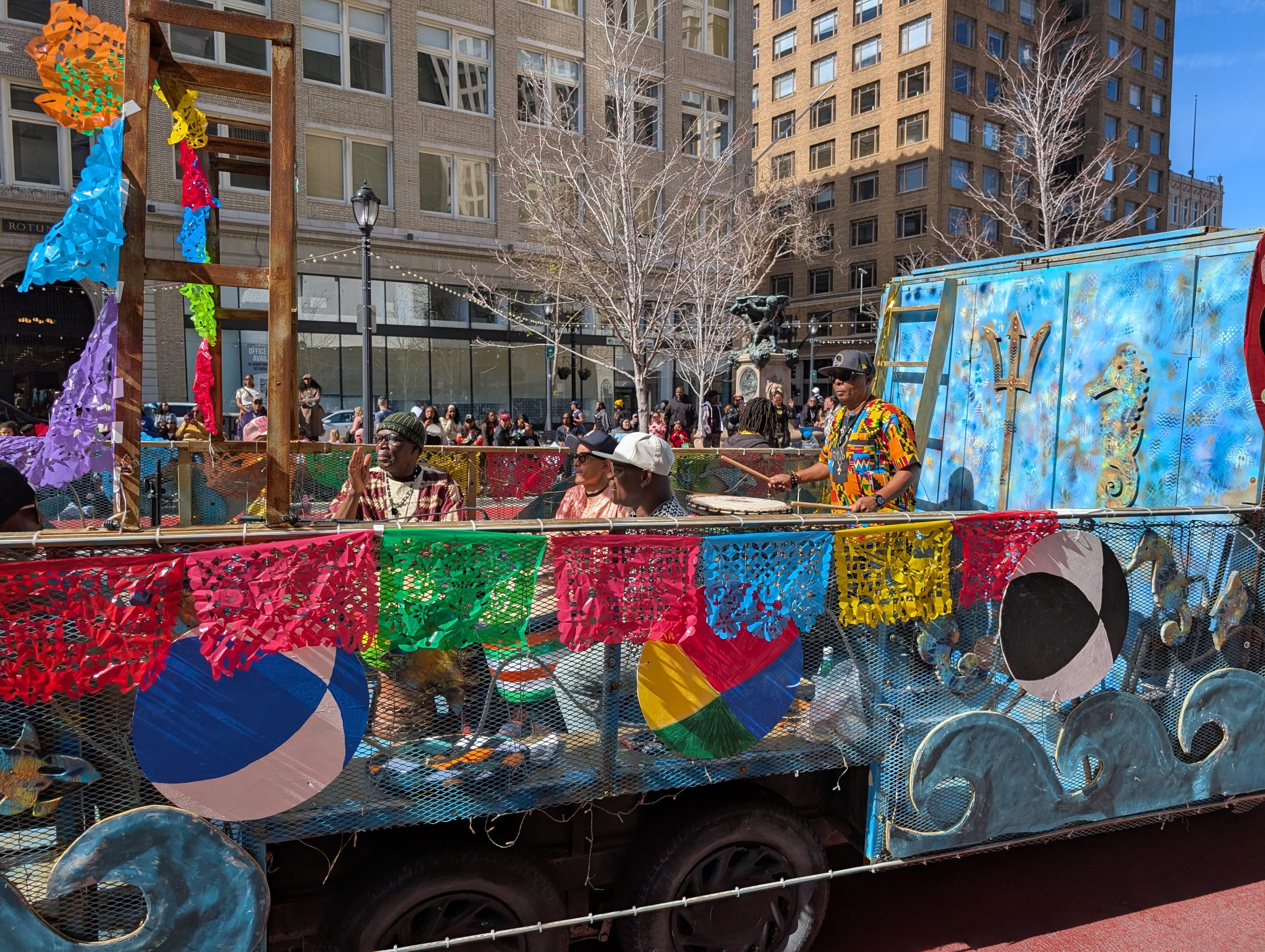 Baba Mosheh wearing a colorful shirt is standing on the back of truck drumming